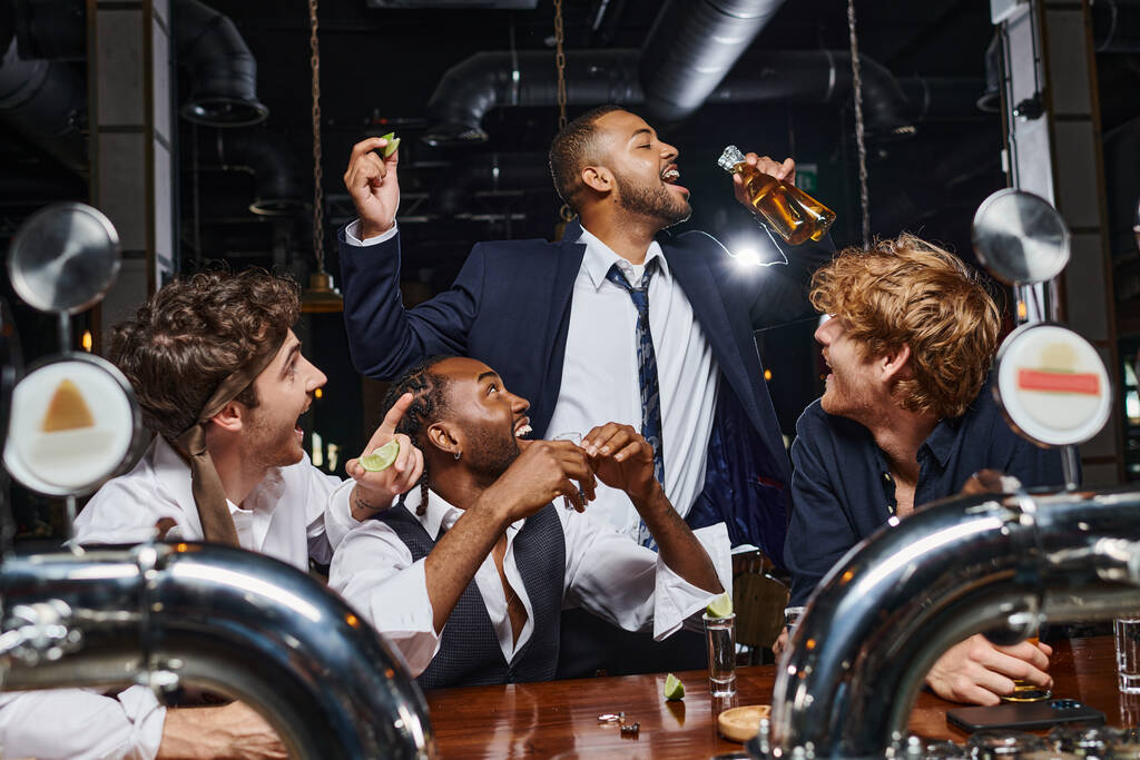 happy men looking at african american friend drinking beer from two bottles after work in bar - Photo, Image