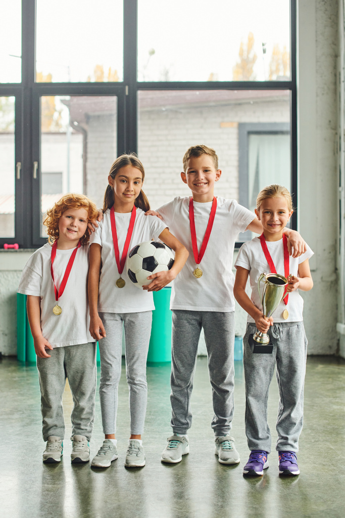 tiro vertical de crianças bonitos pré-adolescentes com medalhas de ouro posando com bola de futebol e troféu - Foto, Imagem
