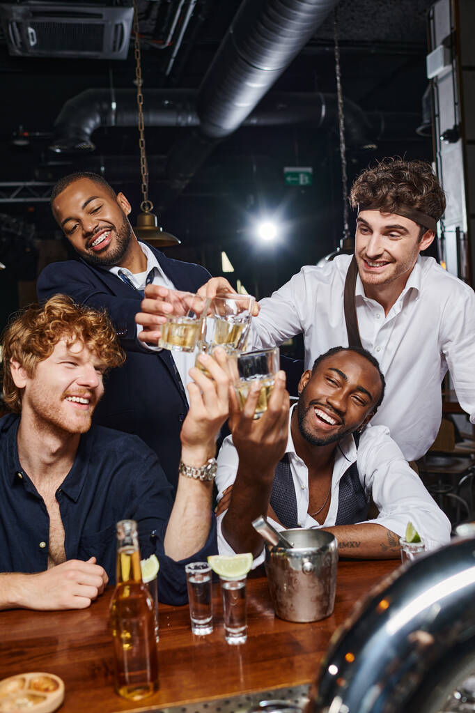 cuatro felices amigos multiculturales brindando con vasos de whisky cerca de tragos de tequila y cerveza en el bar - Foto, imagen