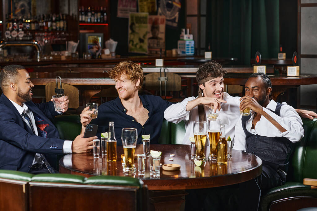 quatro amigos multiculturais felizes bebendo uísque perto de tiros de tequila e cerveja na mesa no bar - Foto, Imagem