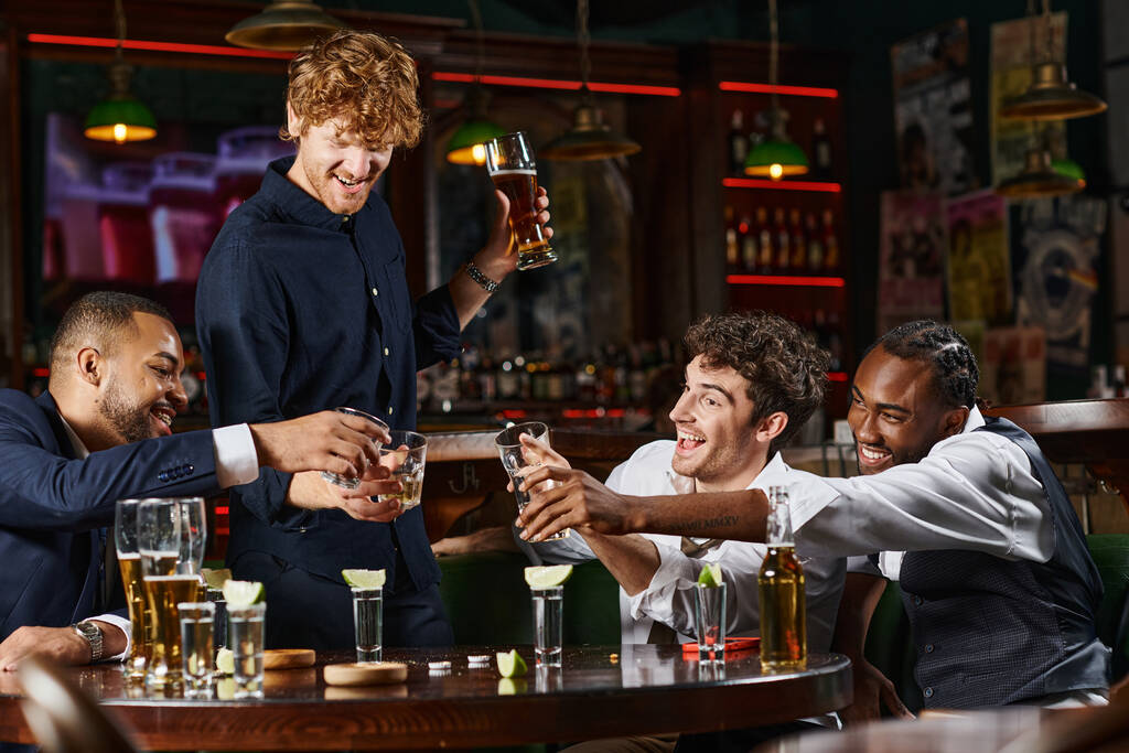Ευτυχισμένοι και μεθυσμένοι πολυεθνικοί συνάδελφοι κάνουν πρόποση με ποτήρια ουίσκι κατά τη διάρκεια του μπάτσελορ πάρτι στο μπαρ - Φωτογραφία, εικόνα