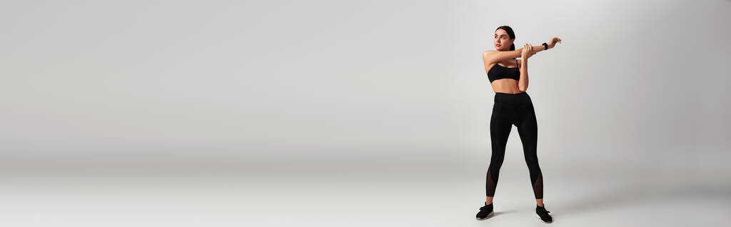 sportowa kobieta w czarnej odzieży sportowej z fitness trackerem na nadgarstku rozciągającym się na szarym tle, baner - Zdjęcie, obraz