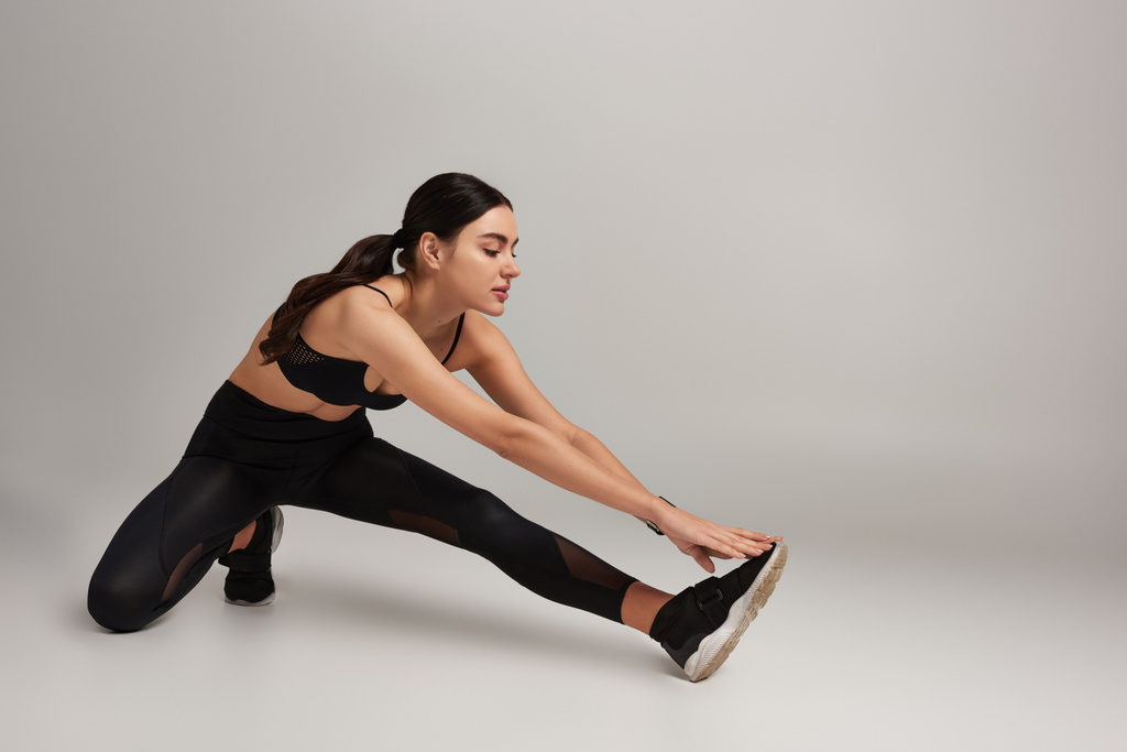 aantrekkelijke vrouw in zwart actieve slijtage met fitness tracker op pols stretching been op grijze achtergrond - Foto, afbeelding