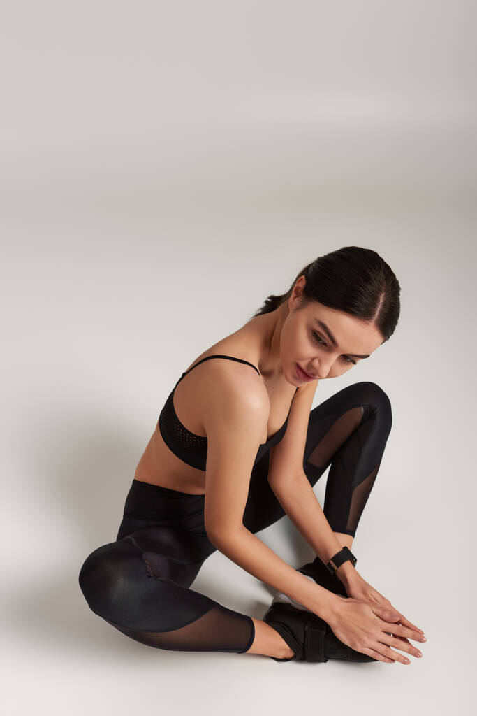 sportiva flessibile in abito attivo nero con fitness tracker al polso seduta su sfondo grigio - Foto, immagini