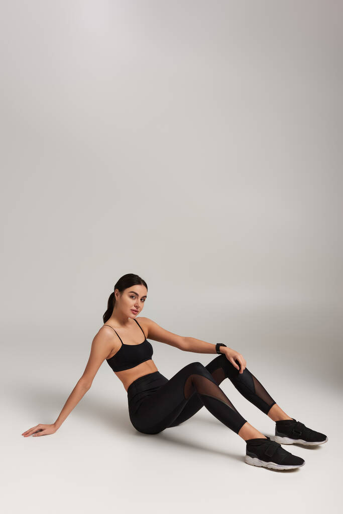 уставшая спортсменка в черной активной одежде с фитнес-трекером на запястье на сером фоне - Фото, изображение