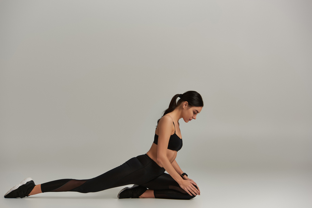 zijaanzicht van flexibele vrouw in actieve kleding en sneakers die zich uitstrekken op een grijze achtergrond, motivatie - Foto, afbeelding