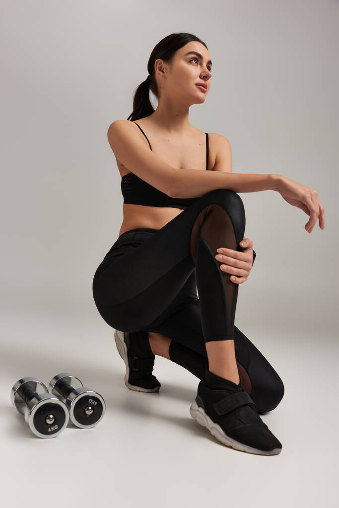 schlanke Sportlerin in aktiver Kleidung, die nach dem Training neben Kurzhanteln auf grauem Hintergrund sitzt - Foto, Bild
