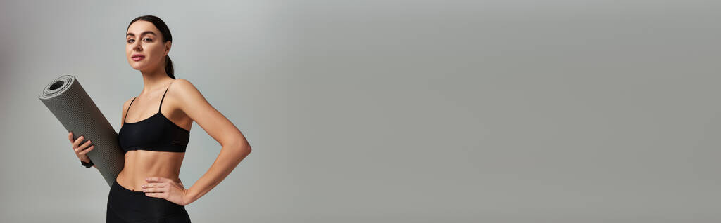 спортсменка в активной одежде держит фитнес-мат и стоит с рукой на бедре на сером, баннер - Фото, изображение
