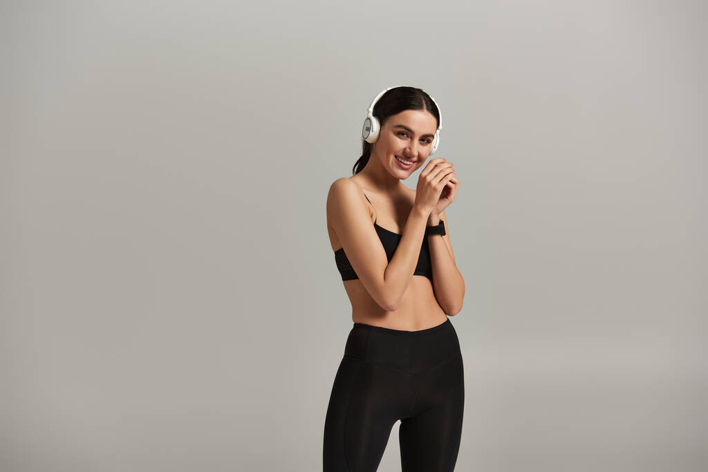 pleased sportswoman in active wear listening music in wireless headphones on grey background - Photo, Image