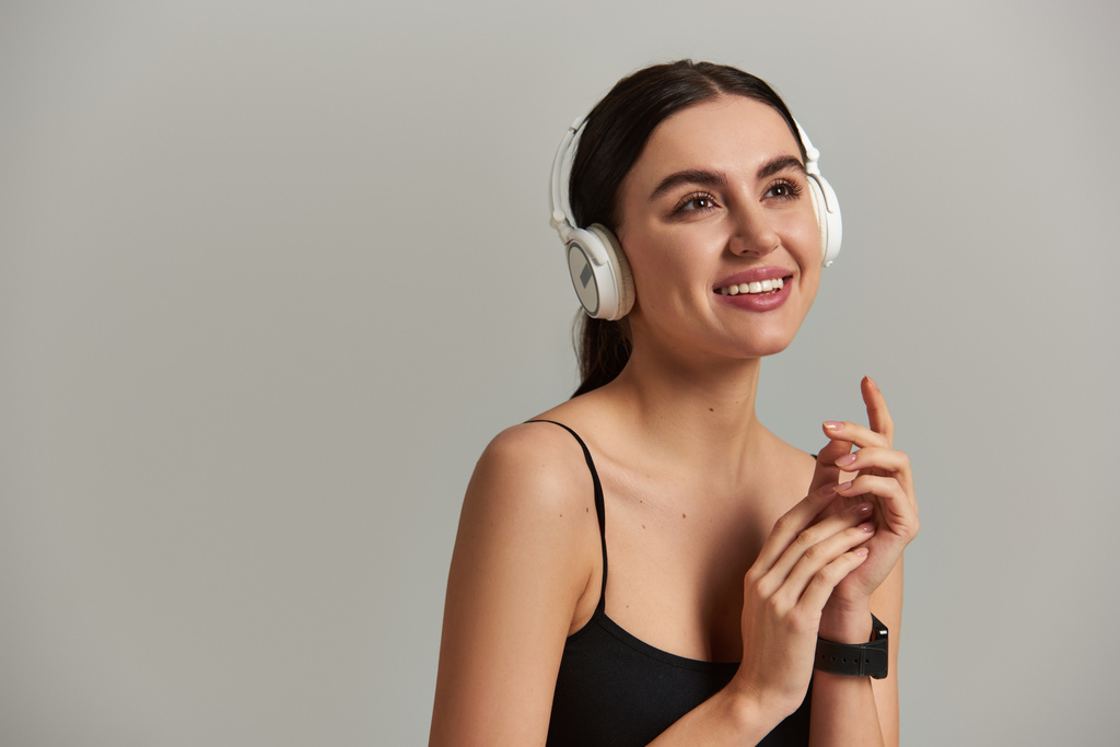 mujer joven feliz en desgaste activo escuchando música en auriculares inalámbricos sobre fondo gris - Foto, Imagen
