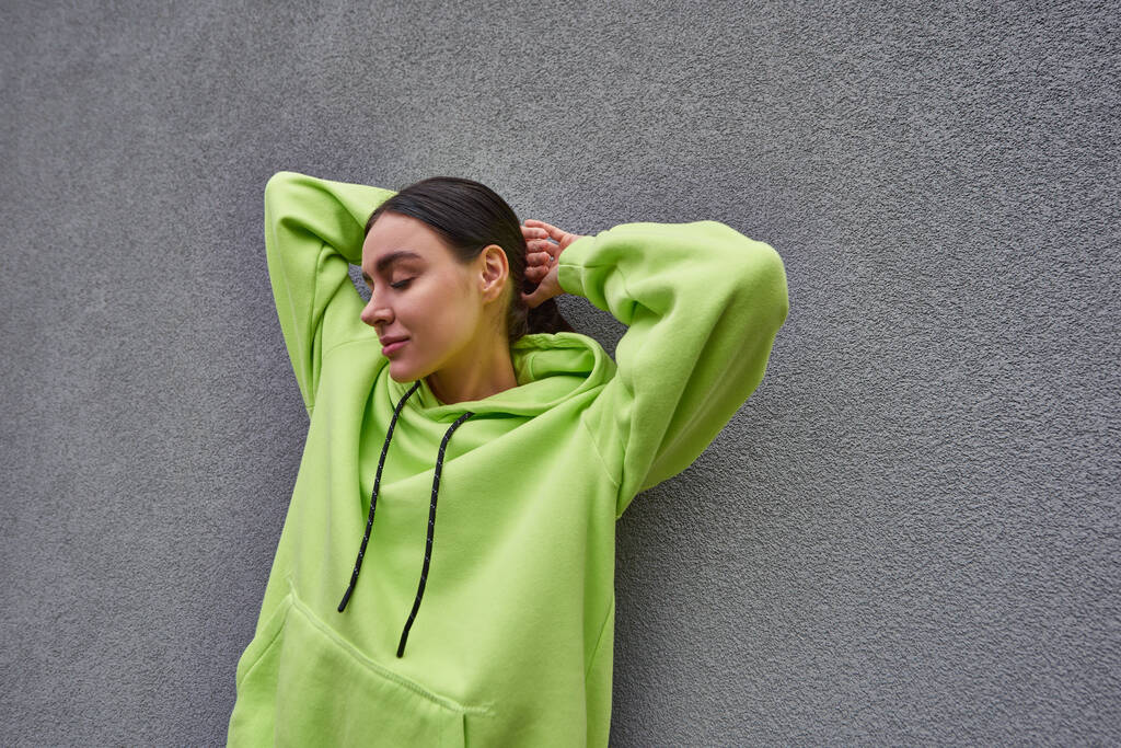 pretty young woman in trendy lime color hoodie posing while standing near grey concrete wall - Photo, Image