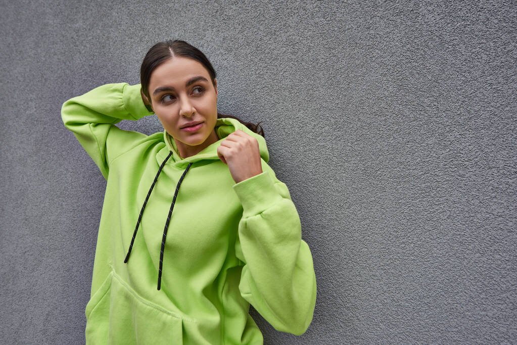 brunette young woman in trendy lime color hoodie posing while standing near grey concrete wall - Photo, Image