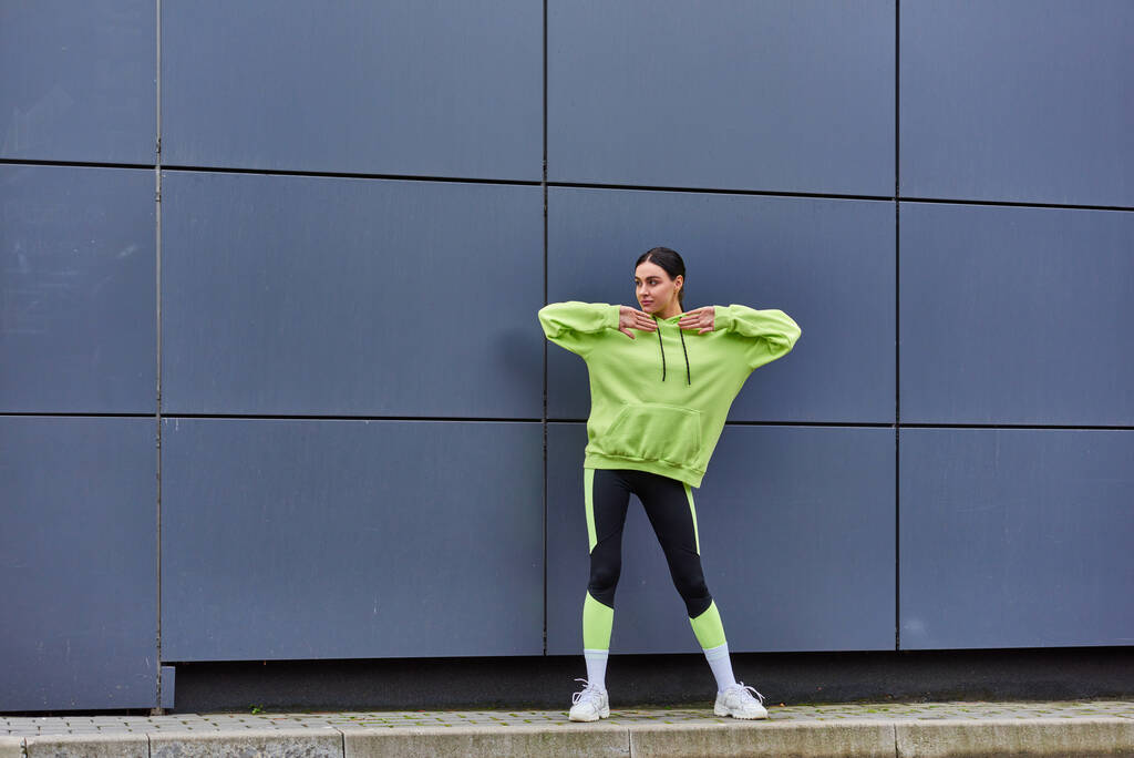 giovane sportiva in felpa con cappuccio e leggings che si scaldano vicino al muro grigio all'aperto, aria fresca e sport - Foto, immagini