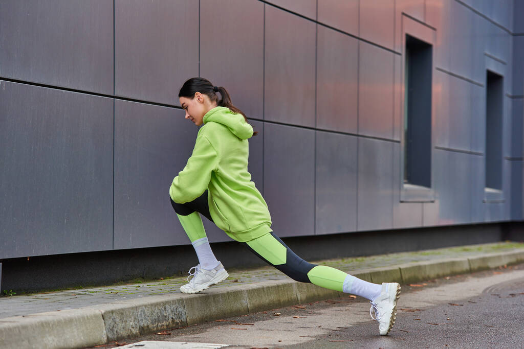 atlética corredora en sudadera con capucha y polainas haciendo embestidas cerca de la pared gris al aire libre, motivación - Foto, Imagen