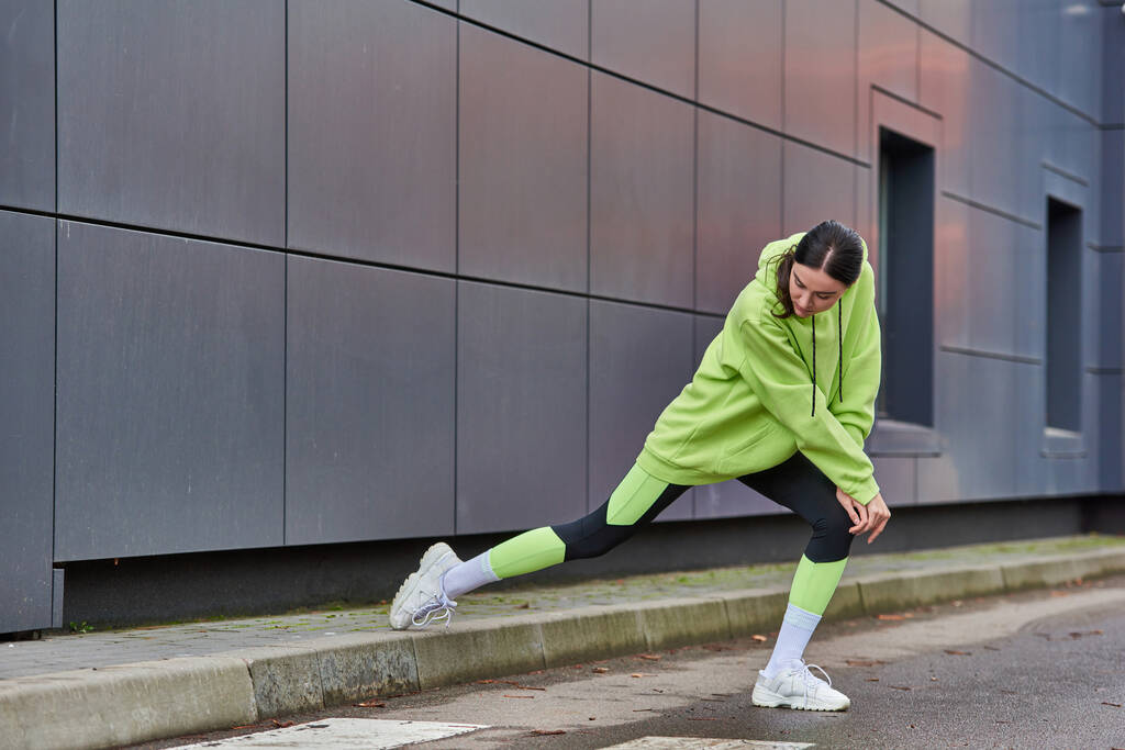 athletic woman in lime color hoodie and leggings doing lunges near grey wall outdoors, motivation - Photo, Image