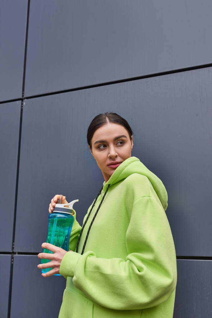 junge Sportlerin in lindfarbenem Kapuzenpulli mit Wasserflasche in der Nähe der grauen Wand im Freien, schaut weg - Foto, Bild