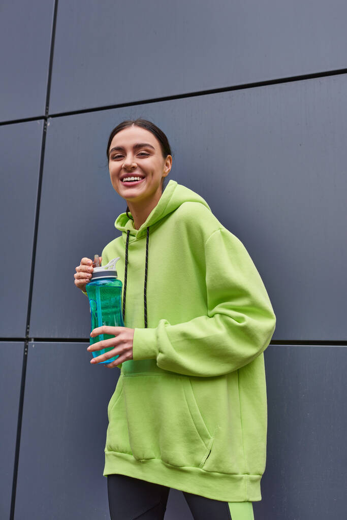 χαρούμενη και νέα αθλήτρια σε λαχανί χρώμα hoodie κρατώντας ένα μπουκάλι νερό κοντά σε γκρι τοίχο σε εξωτερικούς χώρους - Φωτογραφία, εικόνα