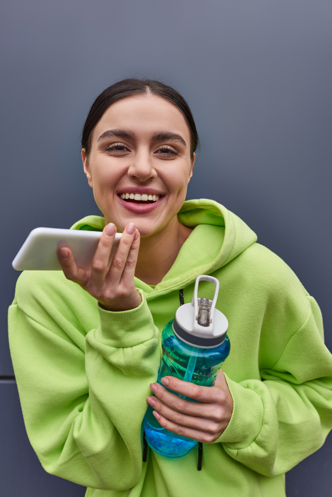 vidám sportos nő kapucnis pulóverben, kezében egy üveg vízzel és hangüzenet rögzítése okostelefonon - Fotó, kép