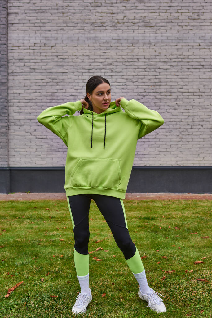 deportista con estilo en sudadera con capucha de color lima y leggings de pie sobre hierba verde, estilo de vida saludable - Foto, imagen