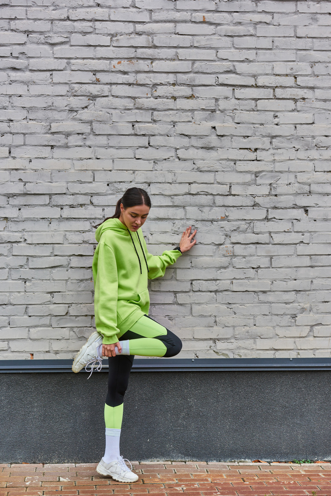 красива молода спортсменка в лаймовому кольорі светр і легінси, що розтягують ногу біля сірої стіни на відкритому повітрі - Фото, зображення