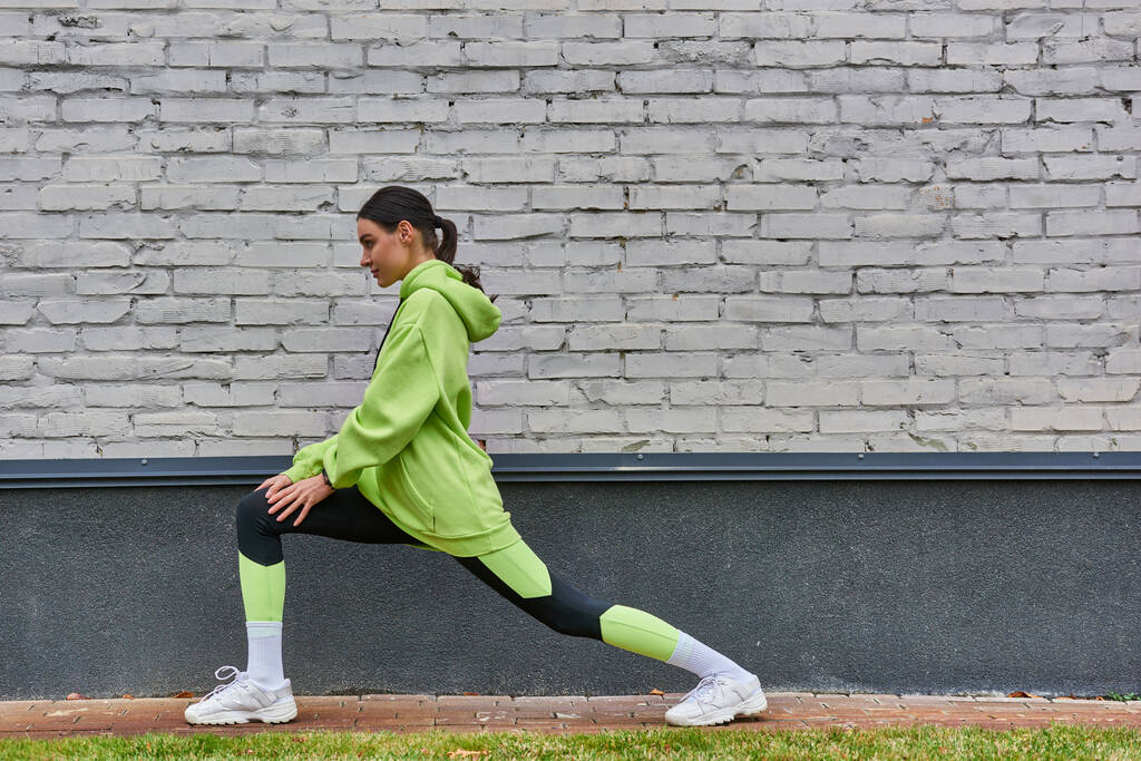 hübsche junge Sportlerin in lindfarbenem Kapuzenpulli und Leggings beim Ausfallschritt vor dem Outdoor-Laufen - Foto, Bild