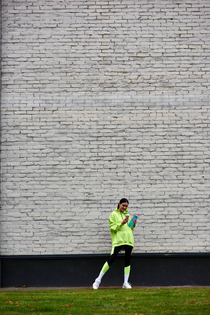 desportista feliz em capuz e leggings usando smartphone e de pé com garrafa com água - Foto, Imagem