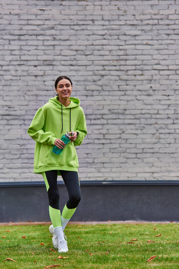позитивная спортсменка в толстовке и леггинсах, держащая смартфон и стоящая с бутылкой с водой - Фото, изображение