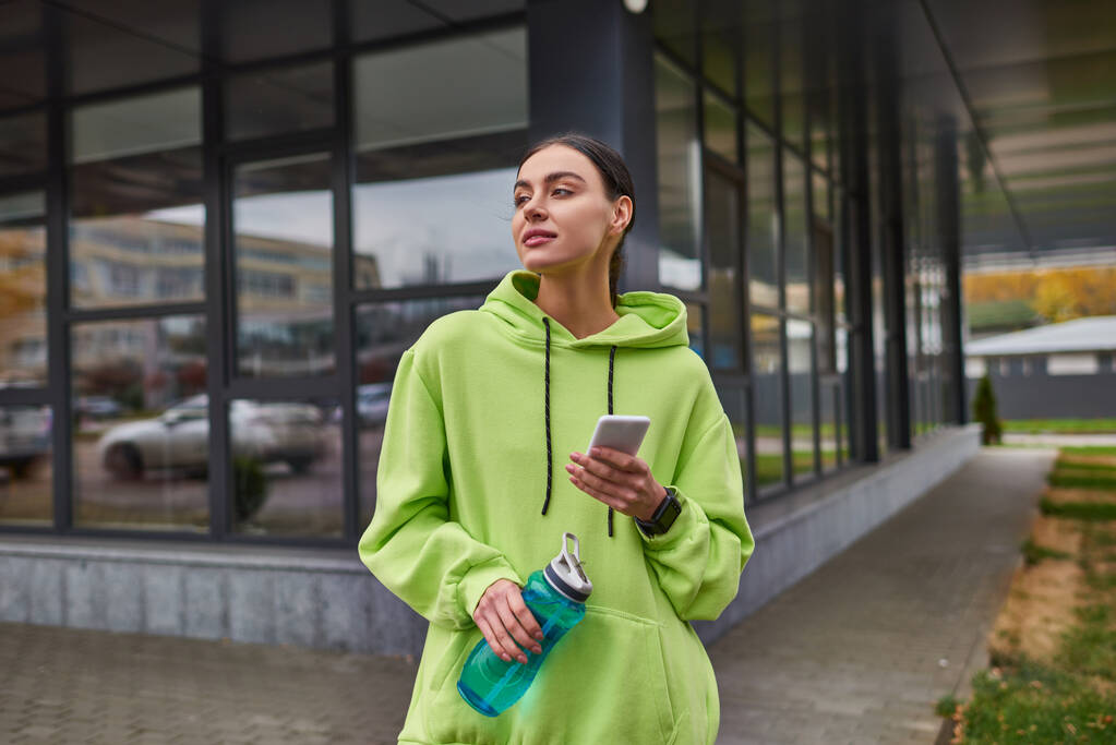 Kireç rengi kapüşonlu spor kadın akıllı telefon kullanıyor ve elinde şişeyle dışarıda bekliyor. - Fotoğraf, Görsel