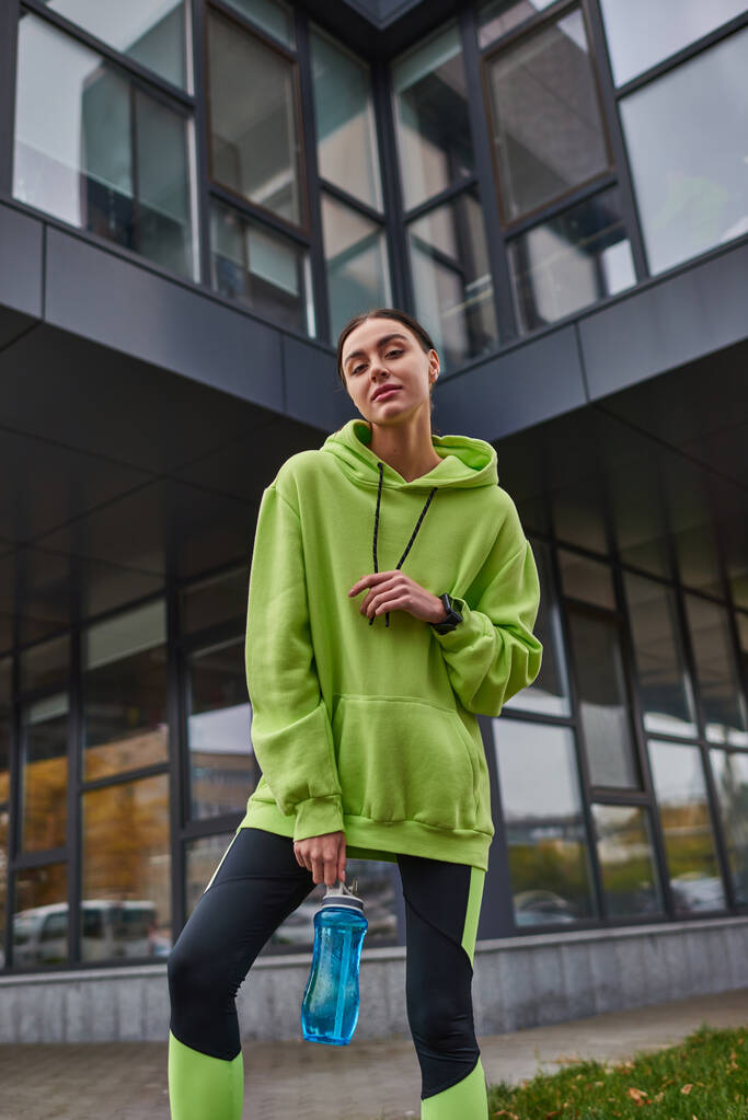 low angle view, sportswoman in oversized hoodie and leggings standing with bottle with water outdoor - Photo, Image