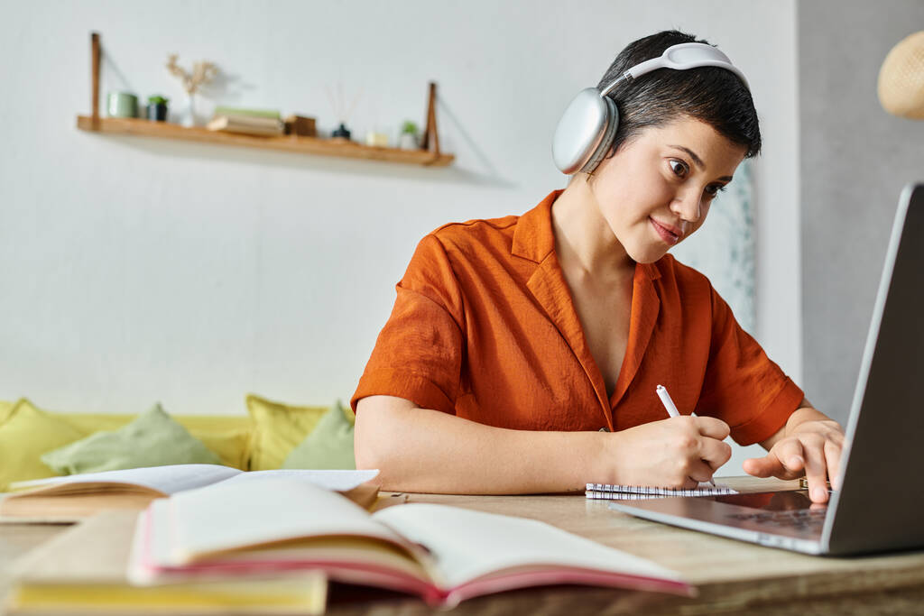 joyful pretty woman in casual outfit with headphones taking notes and looking at laptop, studying - Photo, Image