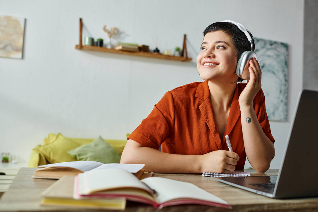 fröhliche junge Studentin in orangefarbenem Hemd, die während ihres Studiums am Laptop Musik hört und wegschaut - Foto, Bild