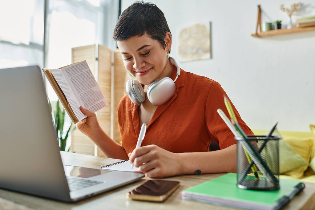 iloinen lyhytkarvainen opiskelija, jolla on kuulokkeet ja kirja kirjoittaa muistiinpanoja, kannettava tietokone pöydällä, opiskelu - Valokuva, kuva