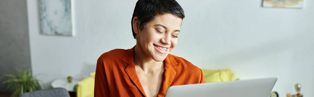 glückliche attraktive Studentin mit Piercing in orangefarbenem Hemd, lächelnd an ihrem Laptop, Bildung, Banner - Foto, Bild