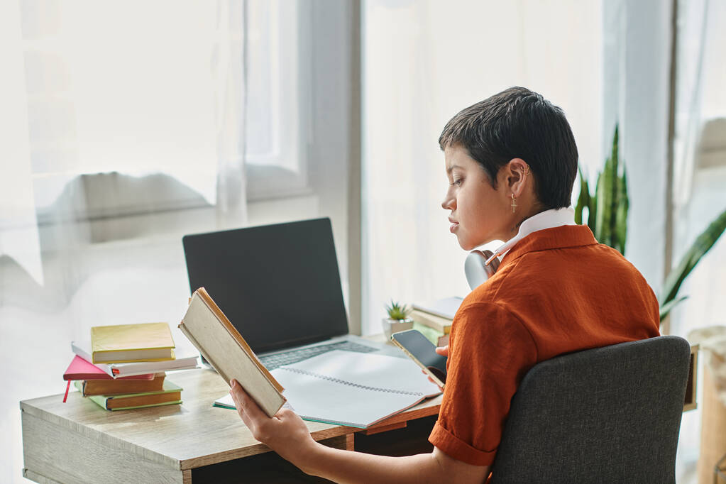 fokussierter Student mit kurzen Haaren, der Lehrbuch mit Telefon in der Hand und Kopfhörer liest, Bildung - Foto, Bild