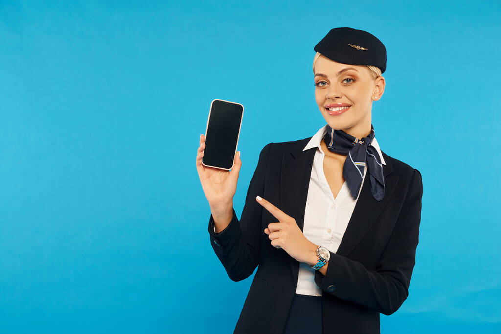 elegant air hostess in stylish uniform pointing at smartphone with blank screen on cyan backdrop - Photo, Image