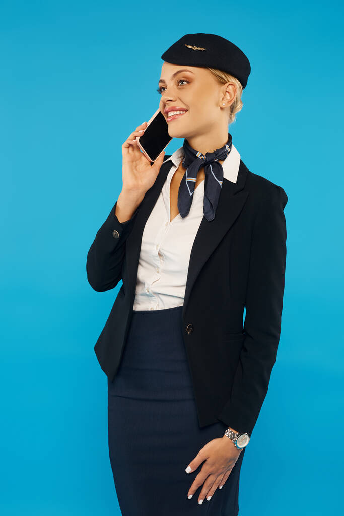 szczęśliwa i elegancka stewardessa z torbą podróżną rozmawiająca przez telefon komórkowy na niebieskim tle, pełna długość - Zdjęcie, obraz