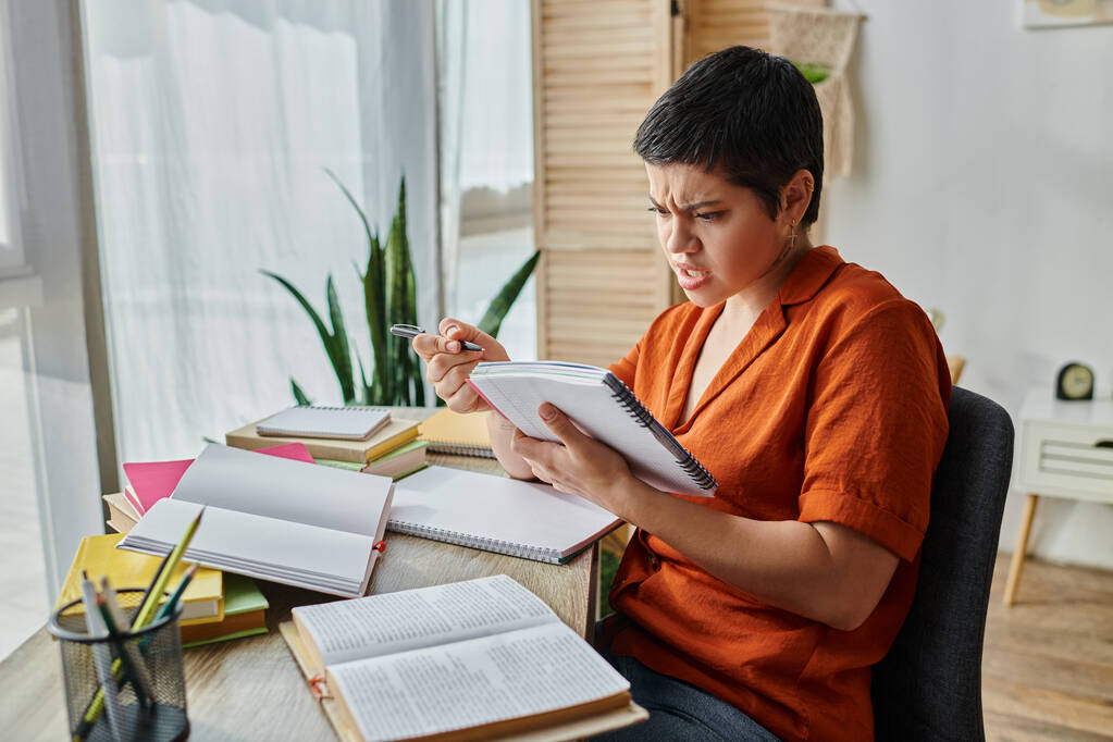 confuso de cabelos curtos estudante do sexo feminino em camisa laranja olhando para o seu livro enquanto estuda duro - Foto, Imagem