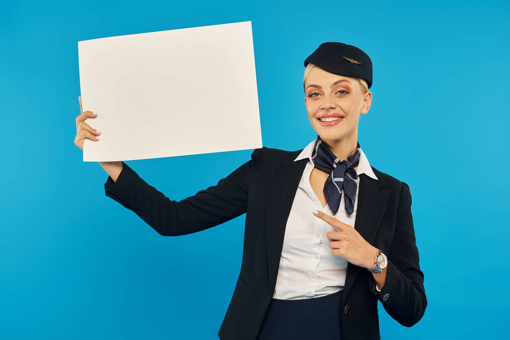 κομψή και χαμογελαστή αεροσυνοδός με στολή που δείχνει με το δάχτυλο σε λευκή αφίσα σε μπλε φόντο - Φωτογραφία, εικόνα