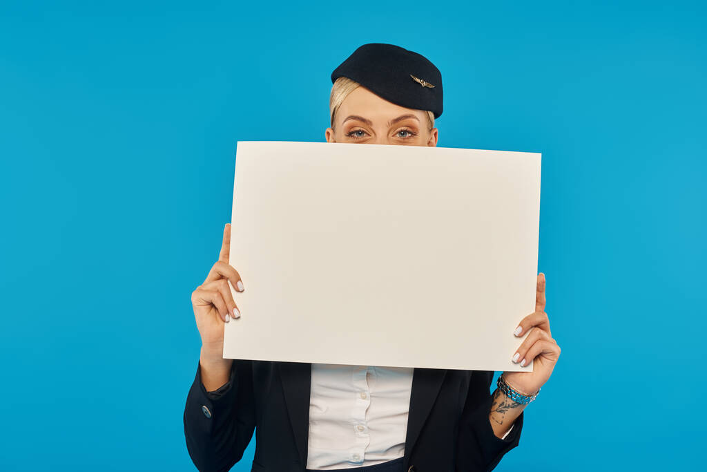 giovane donna in uniforme di assistente di volo oscurante viso con cartello bianco su sfondo blu - Foto, immagini