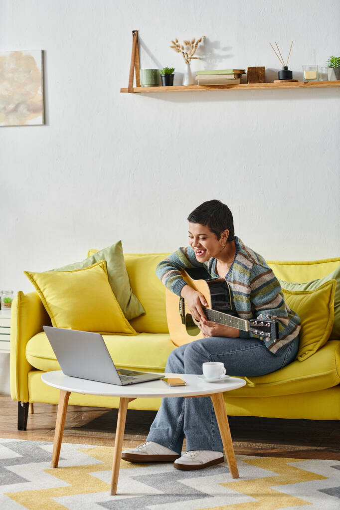 tiro vertical de alegre joven sentada en el sofá con la guitarra mirando en el ordenador portátil en la clase remota - Foto, imagen