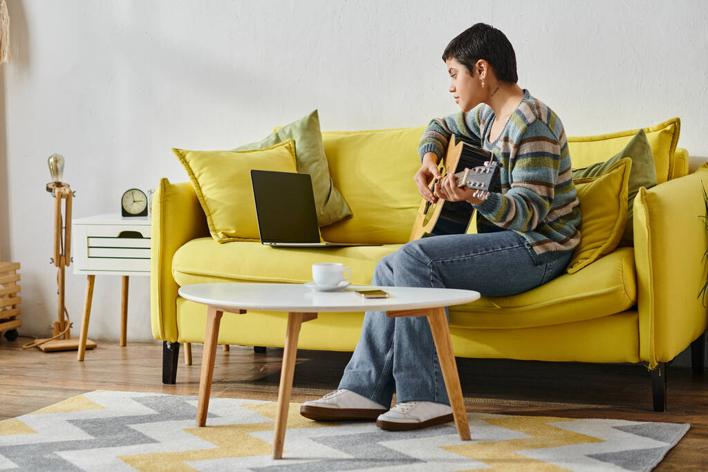красивая молодая женщина в повседневной одежде сидит на диване посещая онлайн урок игры на гитаре, образование - Фото, изображение