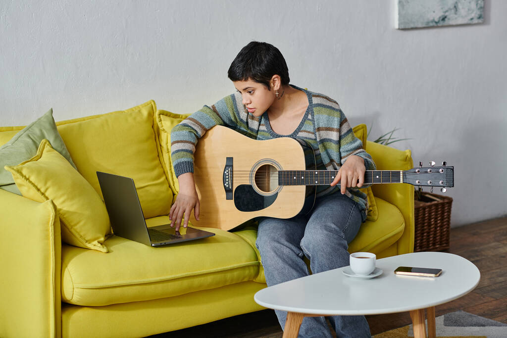 jolie jeune femme assise sur un canapé avec guitare dans les mains et regardant ordinateur portable, éducation à la maison - Photo, image