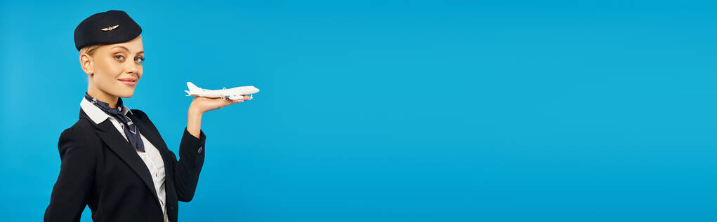 χαρούμενη κομψή αεροσυνοδός σε unform κρατώντας το μοντέλο του αεροπλάνου και χαμογελώντας στην κάμερα σε μπλε, banner - Φωτογραφία, εικόνα