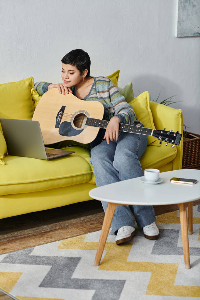 vertikale Aufnahme einer fröhlichen jungen Frau, die mit Gitarre auf dem Sofa sitzt, während sie ferngesteuerten Musikunterricht nimmt, Bildung - Foto, Bild