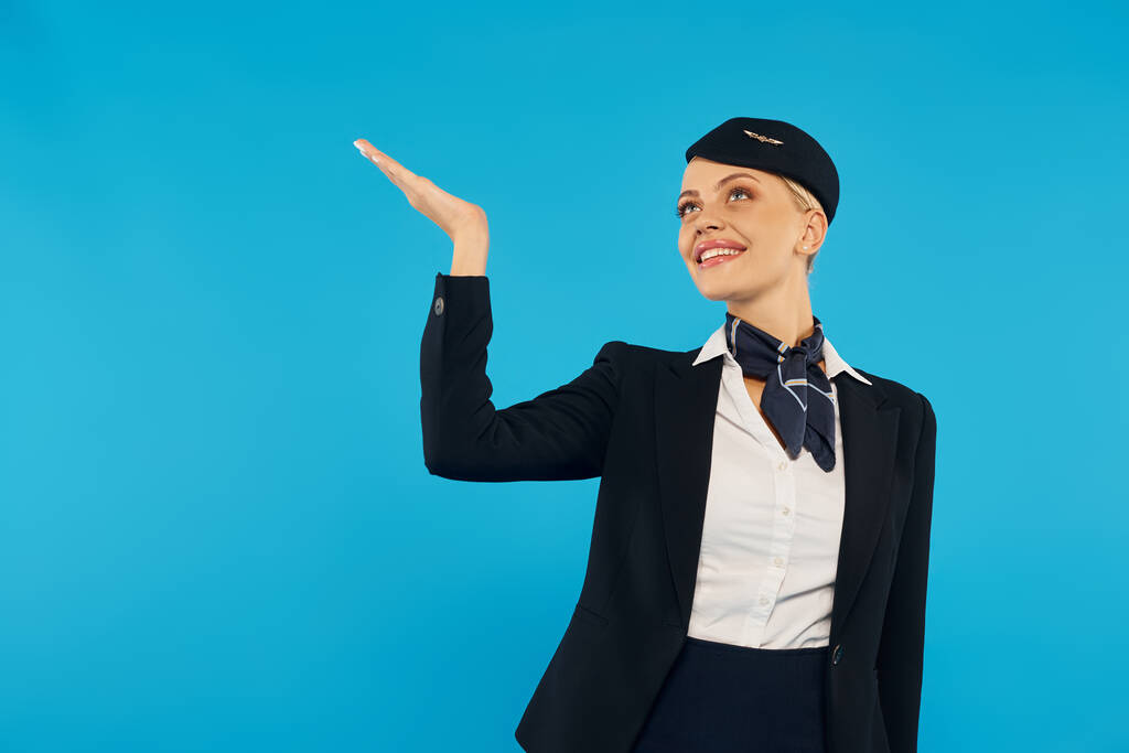 opgewonden en elegant stewardess in corporate uniform kijken omhoog en wijzen met de hand op blauw - Foto, afbeelding