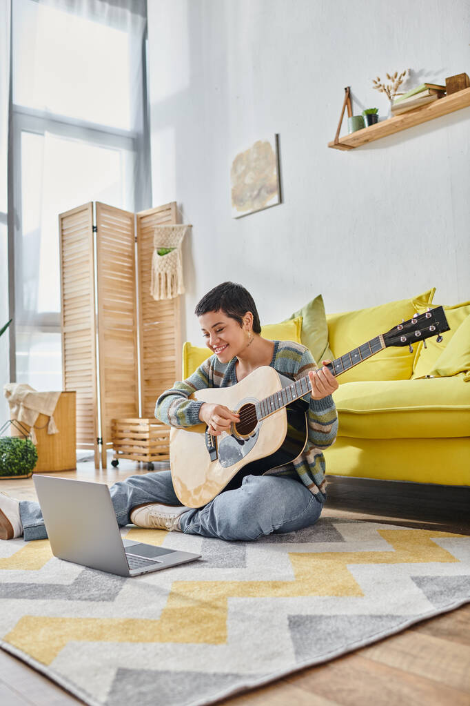 vertikale Aufnahme einer Frau, die auf dem Boden sitzt und mit der Gitarre in die Laptop-Kamera lächelt, Ausbildung zu Hause - Foto, Bild