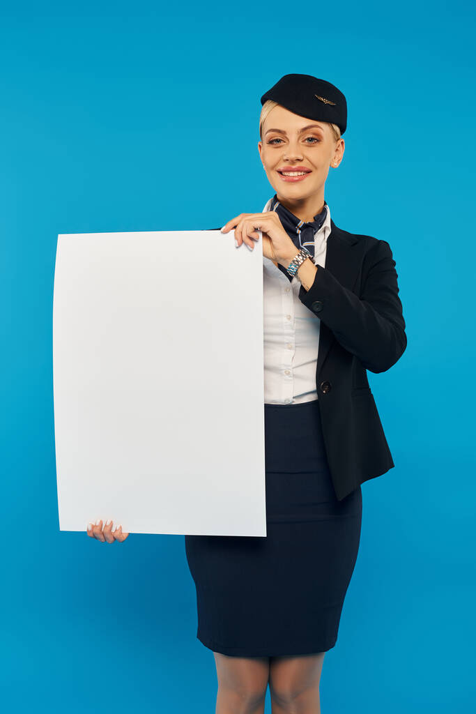 blije lucht gastvrouw in elegante uniform houden blanco bordje en glimlachen op de camera op blauwe achtergrond - Foto, afbeelding