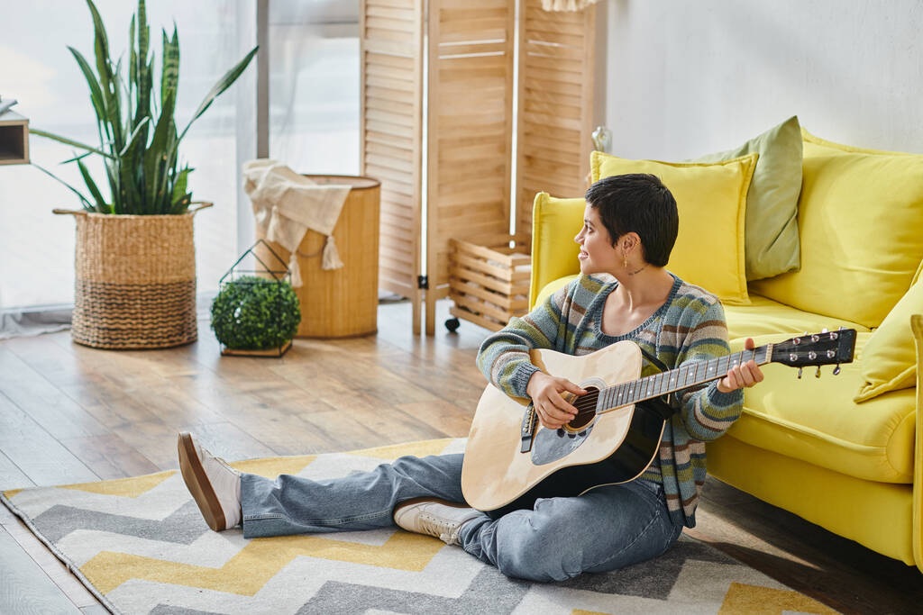 χαρούμενη γυναίκα σε casual ενδυμασία κάθεται στο πάτωμα με κιθάρα και κοιτάζοντας μακριά, εκπαίδευση στο σπίτι - Φωτογραφία, εικόνα