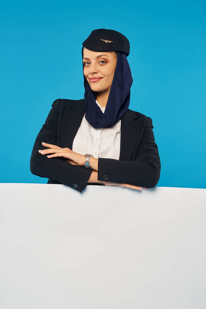 hôtesse de l'air des compagnies aériennes arabes avec les bras croisés souriant à la caméra près de l'affiche vide sur fond bleu - Photo, image