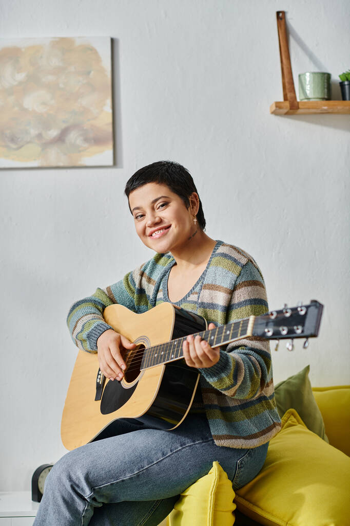 tiro vertical de sonriente mujer feliz en traje casual tocando la guitarra en el sofá, la educación en casa - Foto, imagen